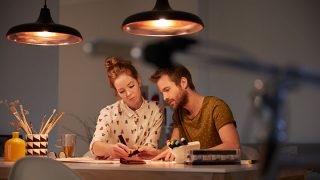 „Stilvolle LED-Pendelleuchten, die ein warmes Licht auf ein zusammen arbeitendes Paar werfen und eine energieeffiziente Beleuchtung für produktive Aufgaben am Abend bieten.“