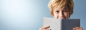 „Junges Kind mit blondem Haar, das hinter einem aufgeschlagenen Buch lächelt und seine Freude am Lesen vor einem hellblauen Hintergrund zum Ausdruck bringt.“