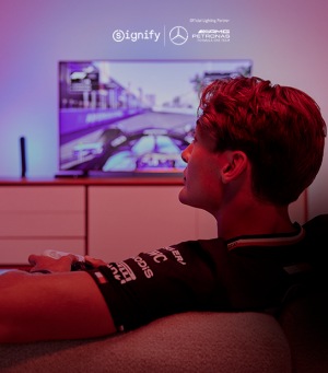 F1 Driver George Russell is sitting on a couch facing the TV while playing a racing video game.