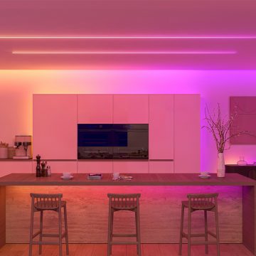 Une cuisine éclairée dans des tons roses et orange vifs grâce à une combinaison de lightstrips dissimulés derrière des moulures du plafond et de lightstrips apparents au plafond. 