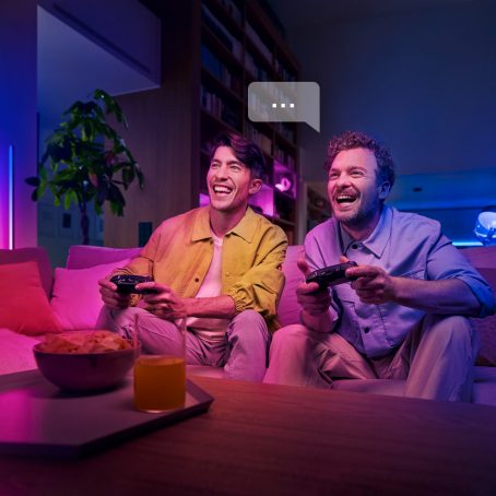 Deux hommes sur le canapé riant en jouant à un jeu vidéo en utilisant leurs voix pour contrôler la boîte de synchronisation hue.