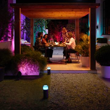 Un groupe d’amis dîne en plein air, entouré d’un éclairage intelligent coloré, réglé sur des nuances de violet et d’orange chaud.