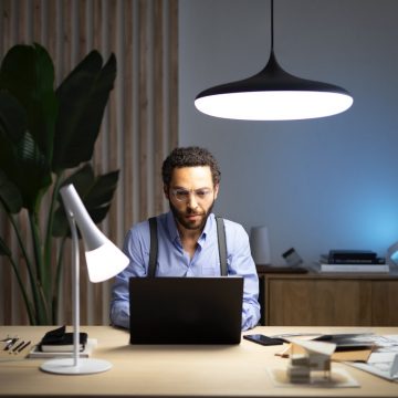 Un homme assis à un bureau devant un ordinateur portable éclairé par une lampe de table et un luminaire suspendu connectés réglés dans des tons blanc froid.
