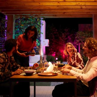Eine Gruppe von Freunden genießt eine Mahlzeit im Freien auf einer Terrasse, die mit farbenfrohem, intelligentem Licht dekoriert ist.