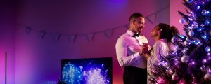 Ein Paar feiert im Wohnzimmer Weihnachten – mit einer Philips Hue LED Weihnachtsbeleuchtung und Lichtern für den Weihnachtsbaum, die den Raum mit smartem blauem und violetten Licht in vielen Nuancen füllen. 