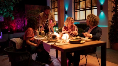 En grupp vänner njuter av en middag utomhus upplyst med färggrann smart utomhusbelysning som är tillgänglig i kampanjen för utomhusbelysning. 