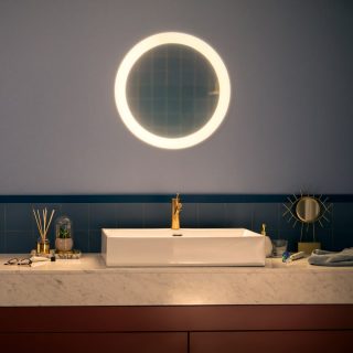 Round smart bathroom mirror with integrated lighting mounted above a sink, illuminating the countertop and faucet area.