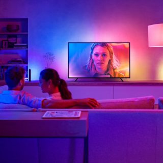 A group sits on a sofa watching TV in a living room, with colorful smart lights illuminating the wall behind the screen.