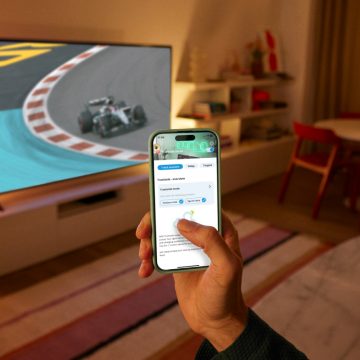 En hand som håller i en telefon som använder WiZ-appen. En TV som visar ett lopp upplyst av WiZ Smart Belysning i bakgrunden.