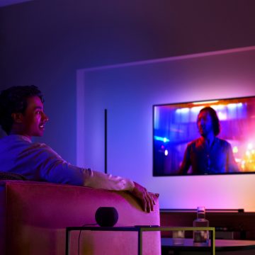 Mann auf einem Sofa vor einem Fernseher mit Surround-Beleuchtung und einem smarten Lautsprecher auf dem Beistelltisch neben ihm
