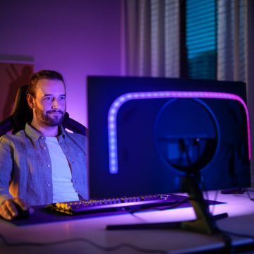 A man sits at a PC monitor fitted with a Hue gradient lightstrip glowing in blue and purple tones of light.