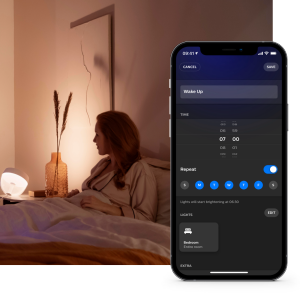 Person in bed using smartphone app to set wake-up light schedule, with smart lamp providing gentle illumination in bedroom.