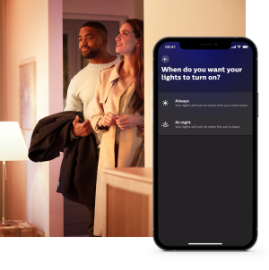 Two people enter a warmly lit home as a smart lighting app displays options for automatic light activation on a smartphone.