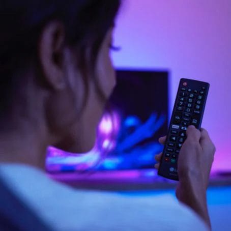 Women pressing a button on her TV remote with colorful TV and lighting in background.