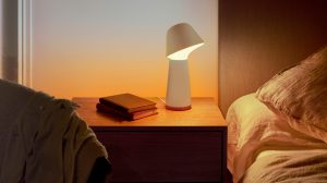Smart table lamp with a white angled shade placed on a wooden nightstand, emitting light onto nearby books and bedding.