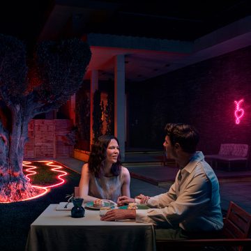 Una coppia cena in un patio, illuminata da strisce di luci al neon per esterni che circondano un albero e un'aiuola e che brillano nei toni del rosa. 