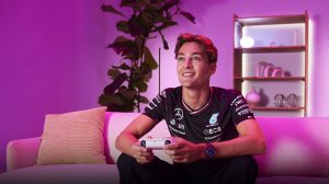 F1-rijder George Russell zit op een bank, houdt een afstandsbediening vast en speelt een videogame in een kamer met roze verlichting.