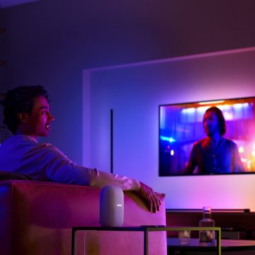 Homem num sofá a ver TV com iluminação envolvente e uma coluna inteligente na mesa de apoio